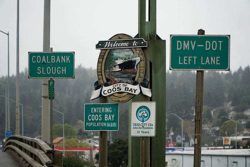 The Welcome To Coos Bay Sign on a rainy day.  It's surrounded by other road signs.