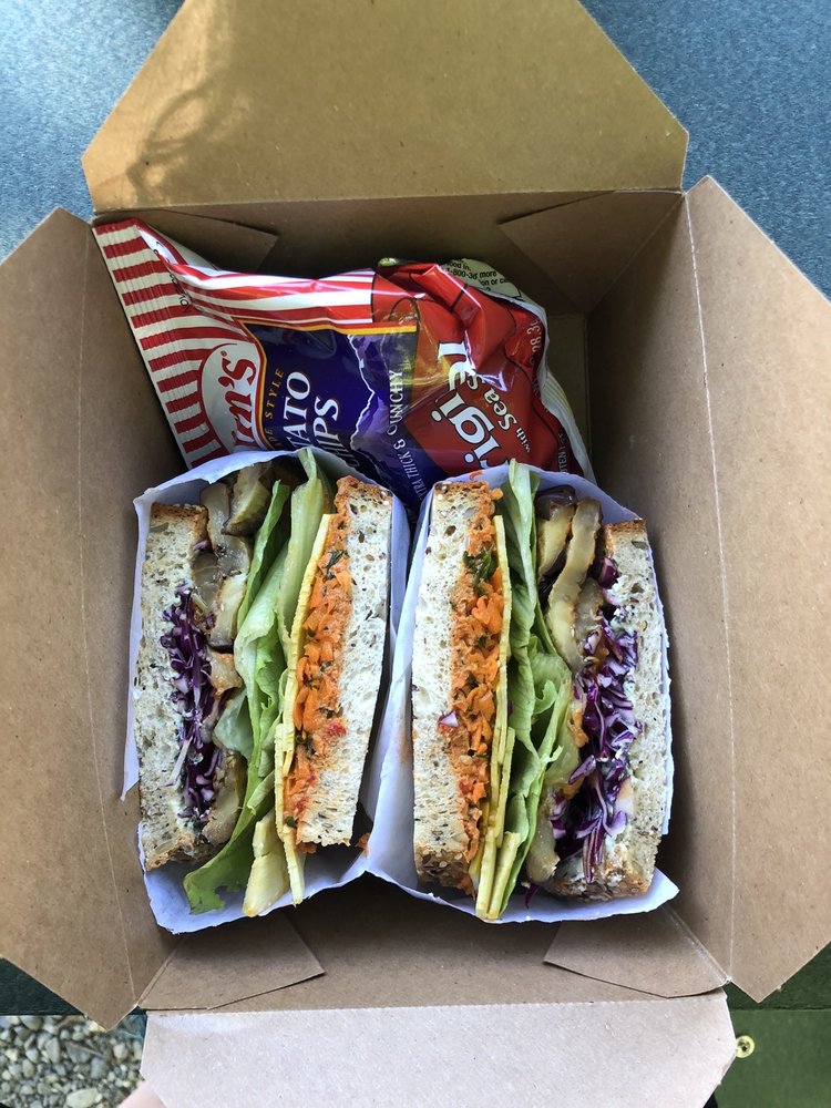 A colorful sandwich and a bag of chips in a cardboard to go box. The sandwich is cut in half and wrapped so it's ready to eat.