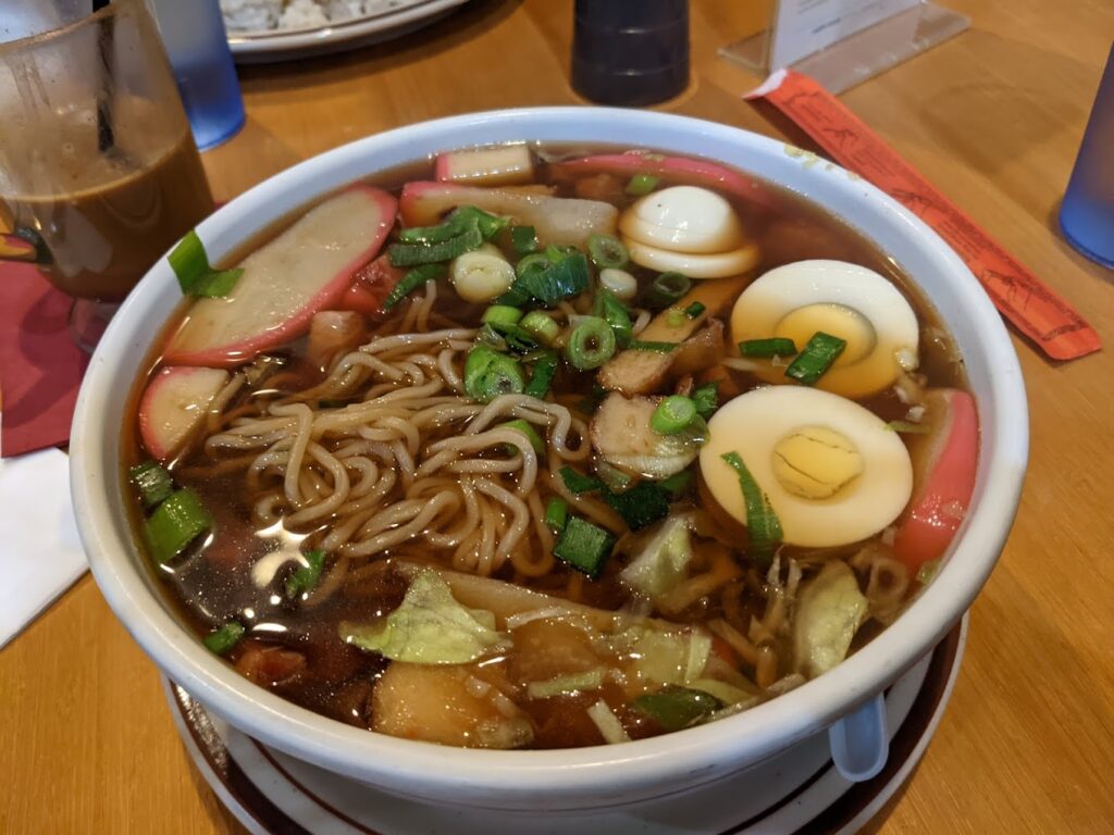 A bowl of broth, noodles, vegetables and boiled egg.