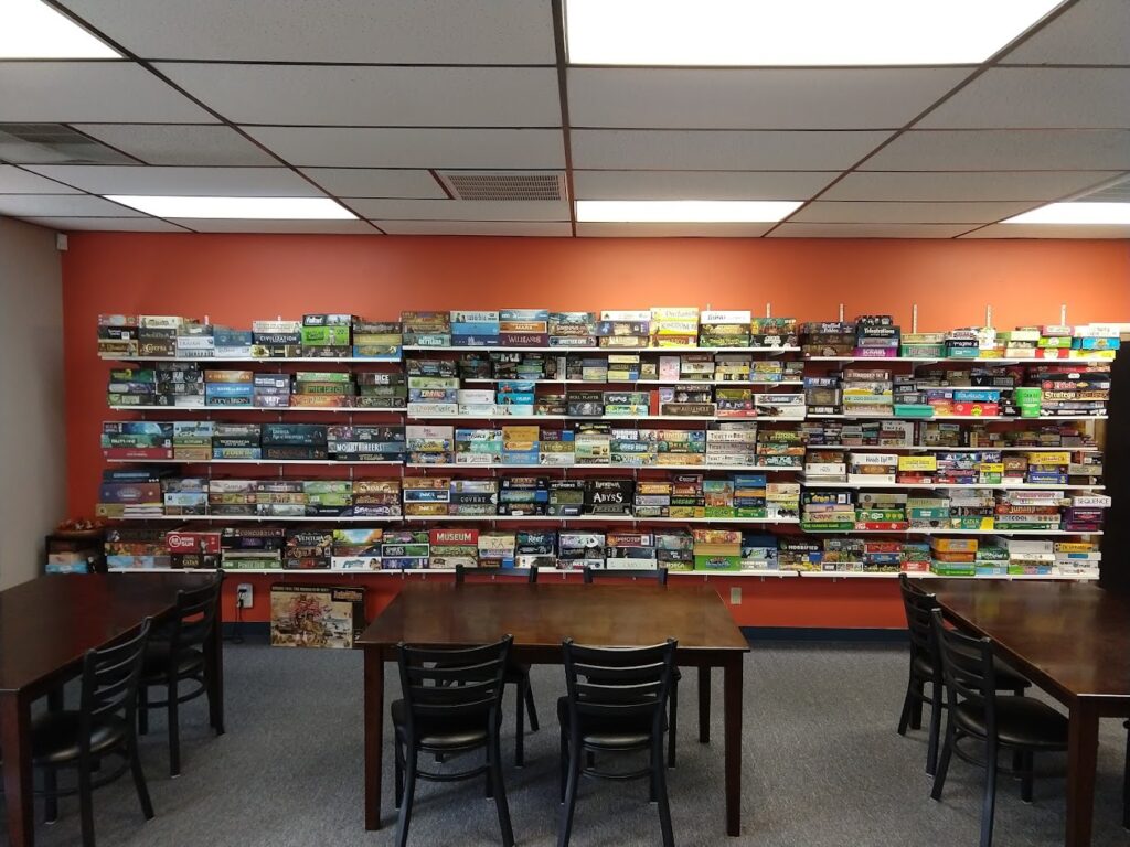 An orange wall covered in shelves of board games. There are wooden tables and chairs to sit at and play games.