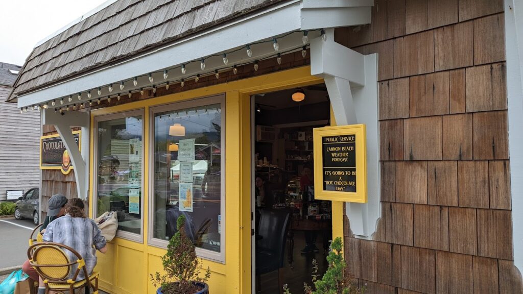 The outside of the Cannon Beach Chocolate Café. It's a building with wood shingles and a dark yellow wall with white trim. It's super cute!