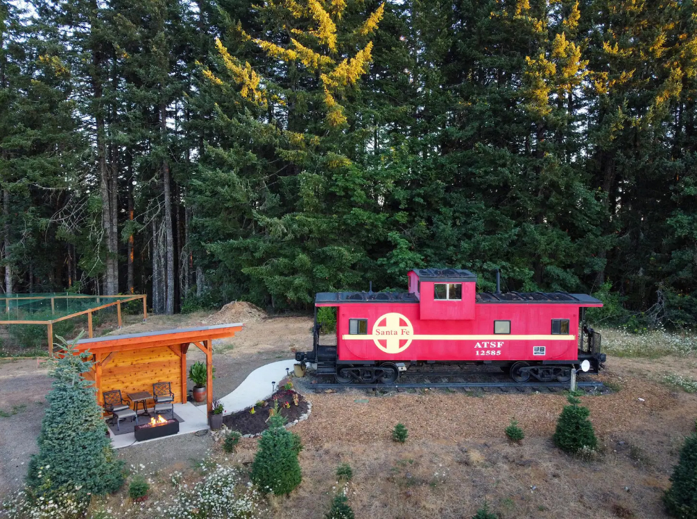 The outside of the bright red train car. There's a paved path leading from the train car to the covered patio with a fire pit.