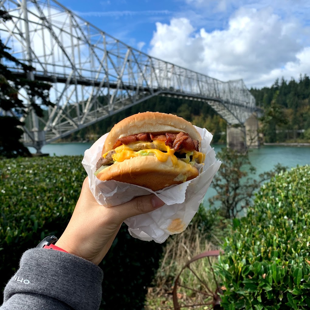 A person holds up a big bacon cheeseburger in front of Bridge Of The Gods.