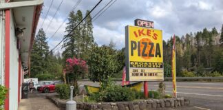 A bright yellow Ike's Pizza sign surrounded by plants.