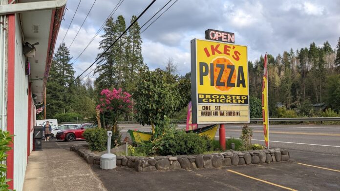 A bright yellow Ike's Pizza sign surrounded by plants.
