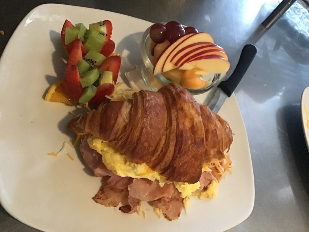 A delicious looking sandwich made out of a croissant, eggs and meat. There is fruit on the plate next to it.