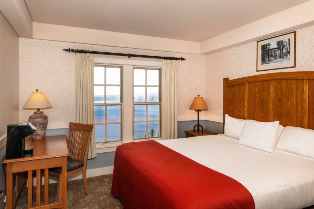 A room in the Crater Lake Lodge. It has a bed with a white blanket and a red blanket, a desk and chair, and a window with white curtains overlooking Crater Lake.
