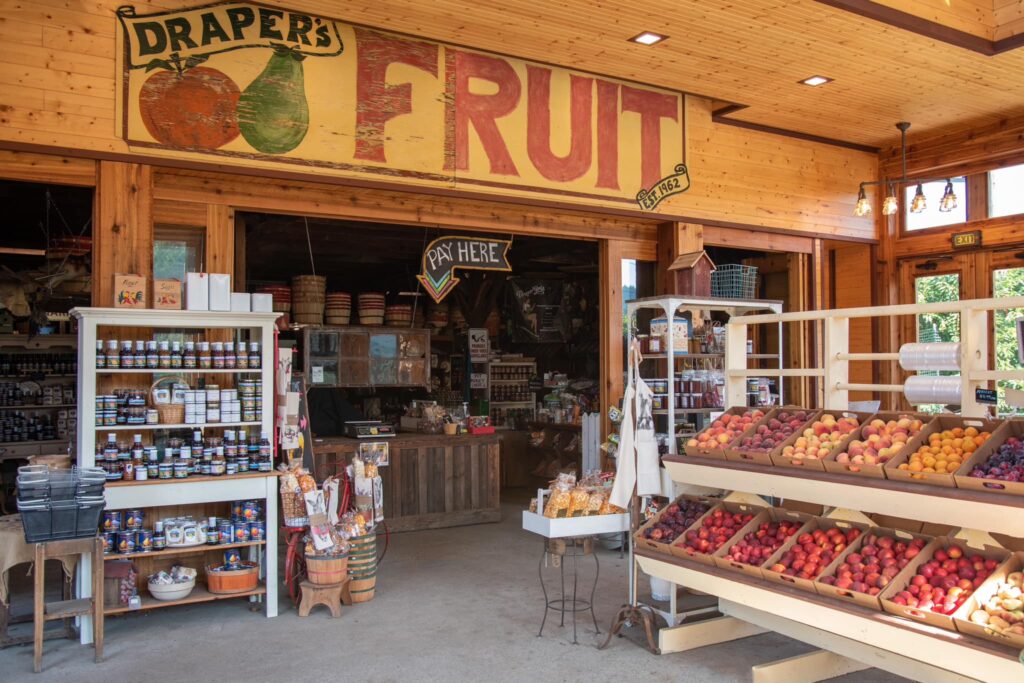 The outside of Draper Girls Country Farm Store. There are boxes of ripe fruit, jars of honey, and a large hand painted wooden sign with the words 'Drapers' and 'Fruit'. It's a really cute little store with wood walls.