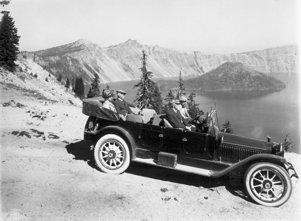 An old black and white photo of a black car with no top on the rim of Crater Lake with several people inside.