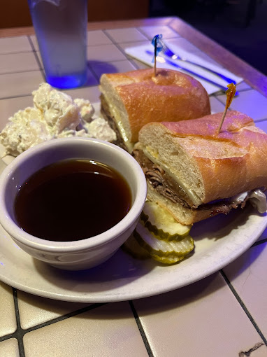 A French Dip sandwich with au jus in a bowl, and sliced pickles on the plate.