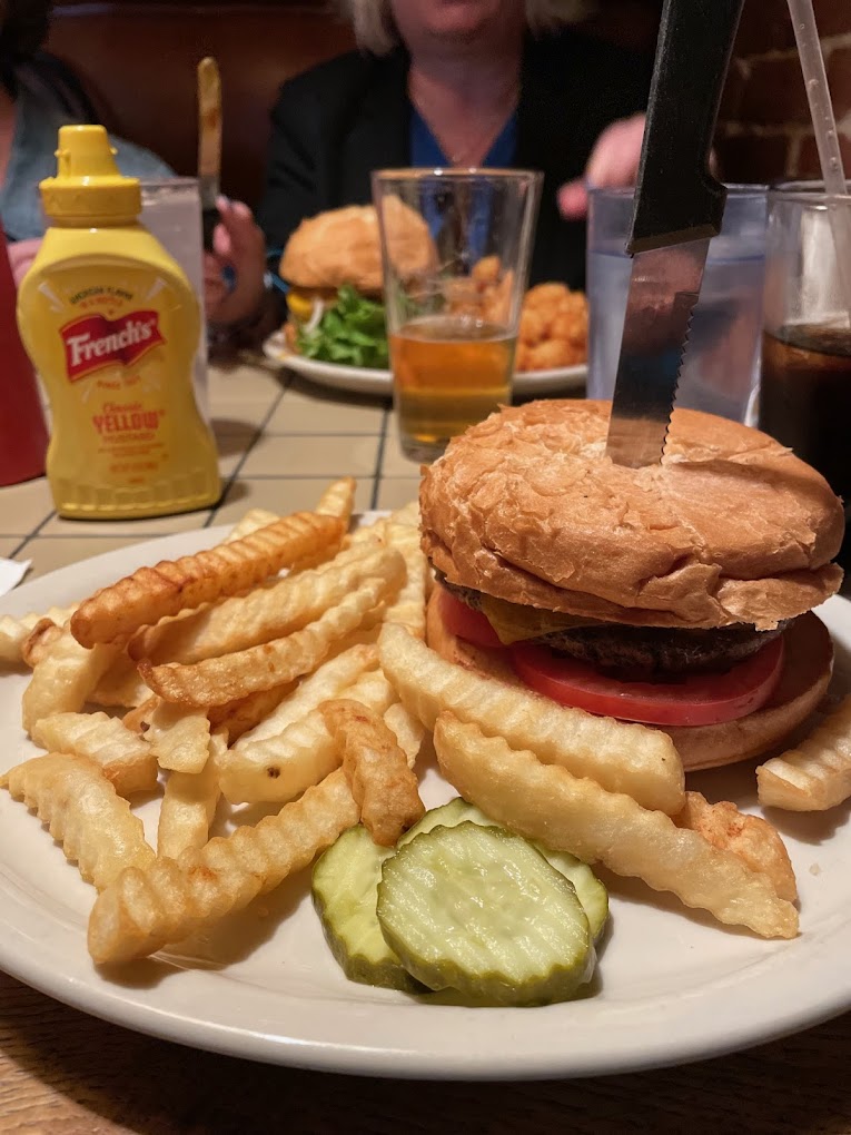 A burger with fries and pickles on the side. There's a large steak knife speared through the top of the burger holding it together.