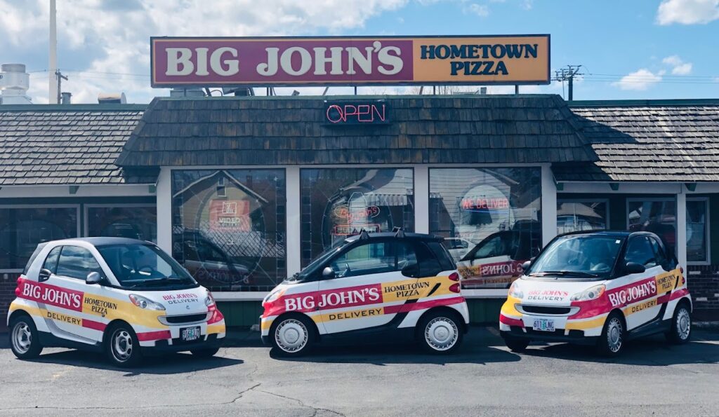 The outside of Big John's Pizza in Pendleton, Oregon. In the photo there are several little white delivery cars for Big John's parked out front.