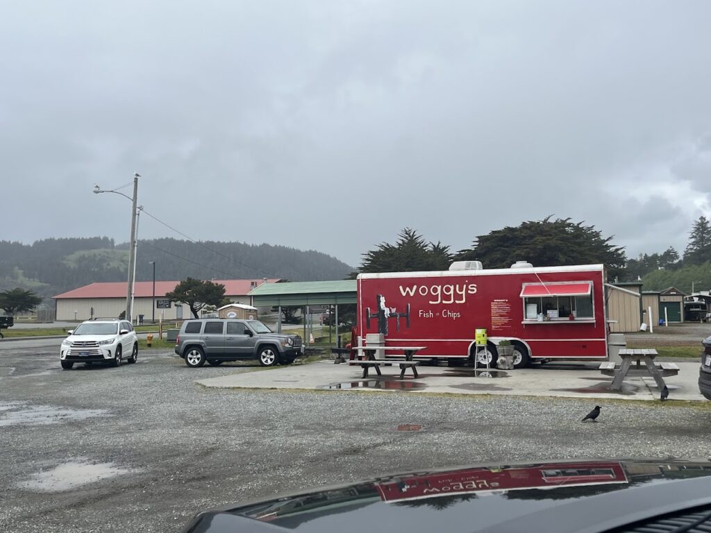 Woggy's food truck. It's red and is sitting in a gravel parking lot on a concrete pad. There are two wooden picnic benches on site to eat at.