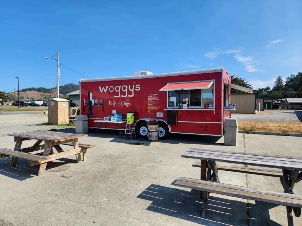 Woggy's food truck. It's red and is sitting in a gravel parking lot on a concrete pad. There are two wooden picnic benches on site to eat at.