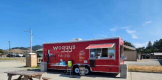Woggy's food truck. It's red and is sitting in a gravel parking lot on a concrete pad. There are two wooden picnic benches on site to eat at.