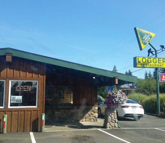 The outside of The Logger Restaurant in Knappa near Astoria, Oregon. The outside is a dark wood with green trim. There's a bright yellow and green sign with a tree and an arrow pointing into the air.