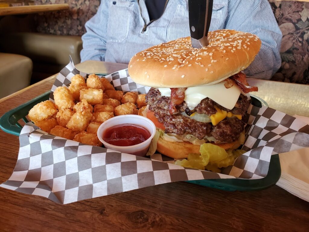 A massive burger with a knife sticking out the top. It has two patties, bacon, Swiss Cheese, American cheese, and other things on it. It's really tall! There are tator tots and ketchup next to it on a black and white checkered paper wrapper.