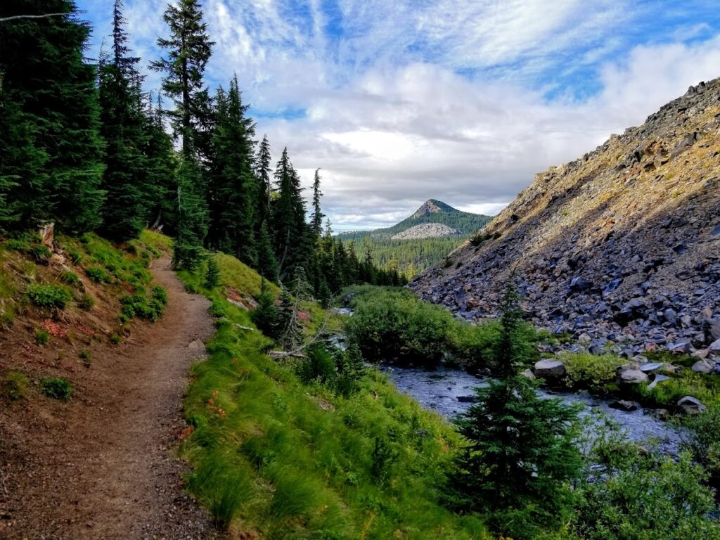 The trail going uphill above the creek. On the left side, there are trees and low shrubs. The creek is on the right side along with a a mountain and rocks from an old lava flow. You can see a mountain peak in the distance.