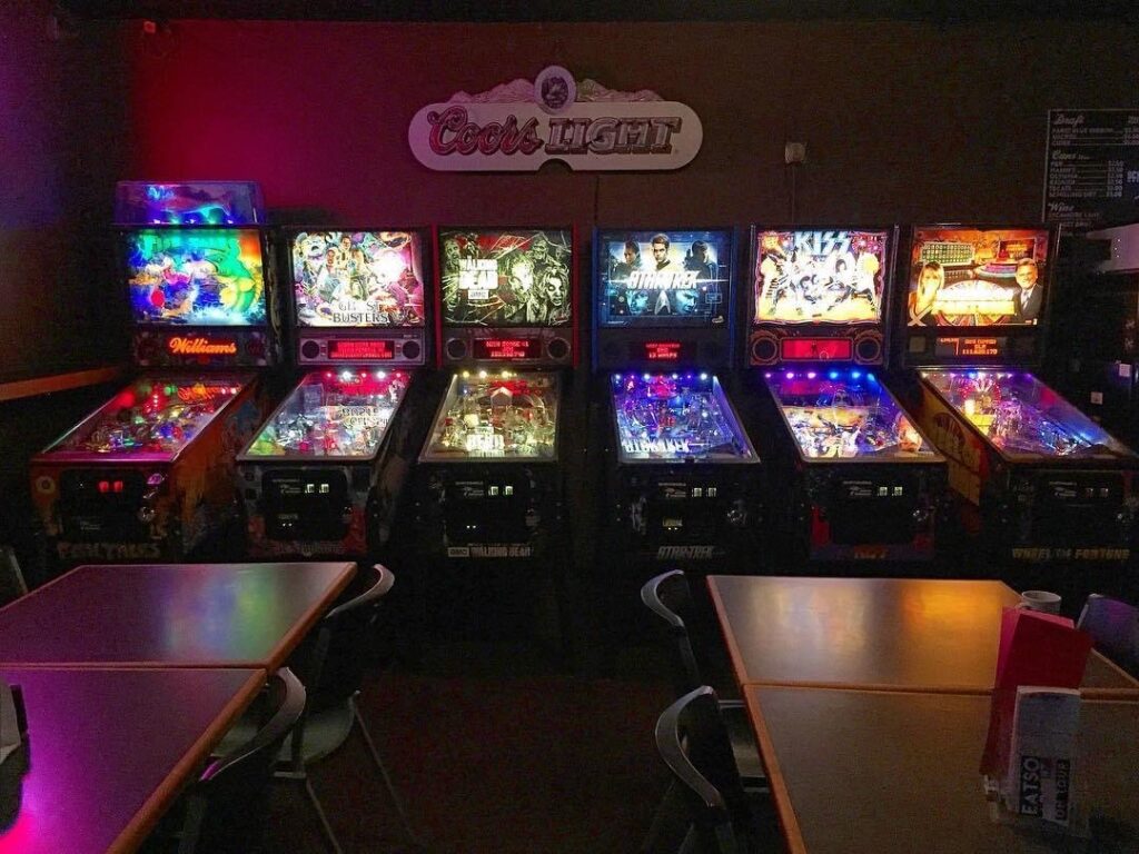 A line of pinball machines all lit up in the dimly lit restaurant.