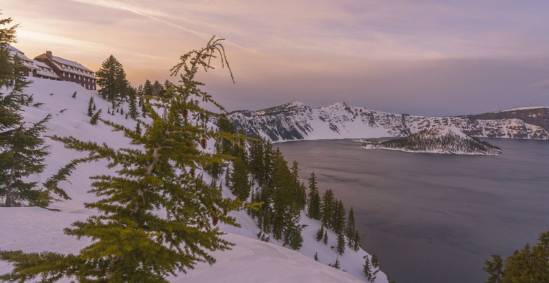 The lodge looks tiny as it sits on a snow covered rim overlooking the massive Crater Lake at sunset.