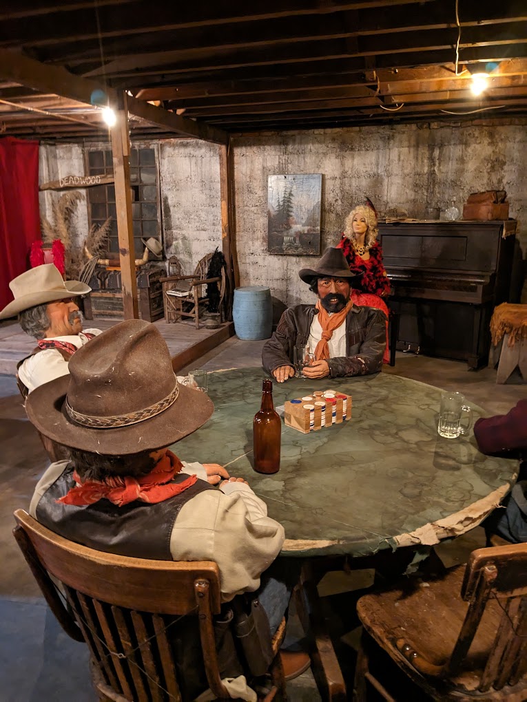Mannequins play cards at a round card table in an under ground room.