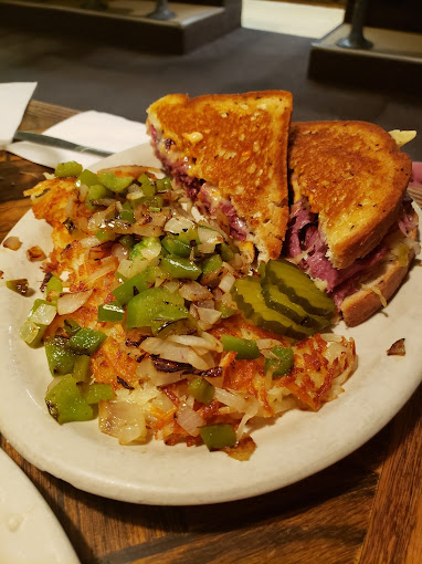 A really delicious looking meat sandwich on toasted bread with hash browns, onions and green peppers. There are sliced pickles on the side as well.