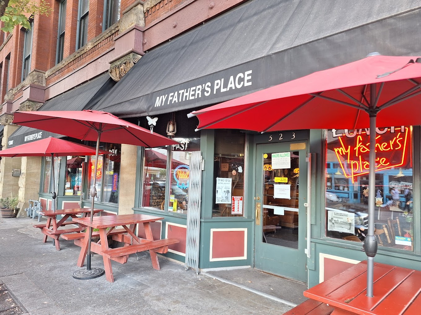 The outside of My Father's Place in Portland, Oregon. It has a black awning with white letters, and there are red picnic tables and red umbrellas out front on the sidewalk.