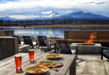 Food sits on outdoor tables by a modern fire pit. In the background you can see a lake, and on the other side of the lake, there's a forest and gorgeous mountains covered in snow. It's a cloudy day.