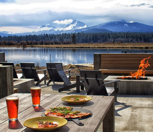 Food sits on outdoor tables by a modern fire pit. In the background you can see a lake, and on the other side of the lake, there's a forest and gorgeous mountains covered in snow. It's a cloudy day.