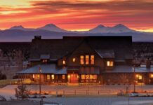 A stunning view of one of the buildings at Brasada Ranch at sunset. There are snowy mountains in the distance, and red orange light coming through wispy clouds.