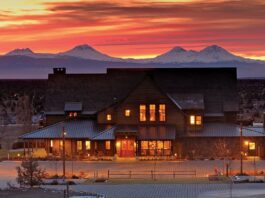 A stunning view of one of the buildings at Brasada Ranch at sunset. There are snowy mountains in the distance, and red orange light coming through wispy clouds.