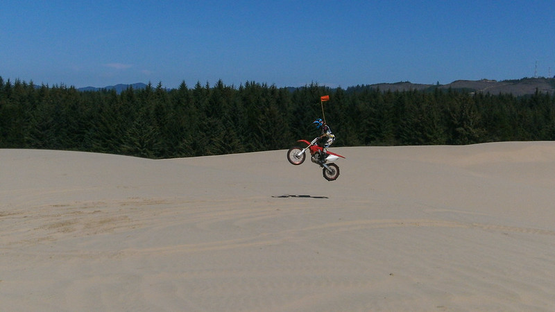 A person on an off road motorcycle jumps into the air at the Oregon Dunes. There is a forest in the background.