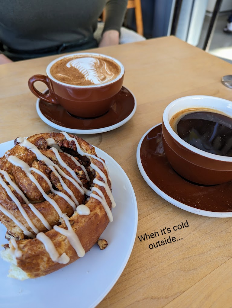 Two mugs of coffee and a huge cinnamon roll with frosting drizzled over the top.