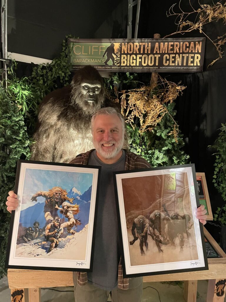 Cliff Barackman holds up two paintings in front of a bigfoot statue at the North American Bigfoot Center.