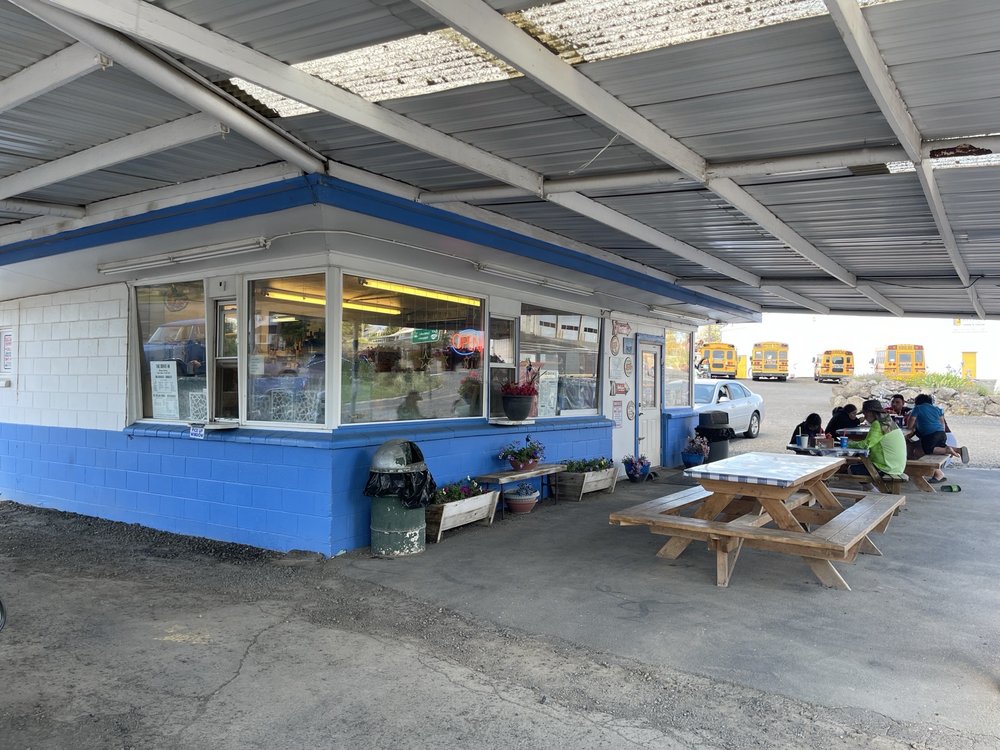 The Drive In as seen from under the overhang. The building is blue and white, and there are picnic tables to eat at underneath the overhang where it's shaded.