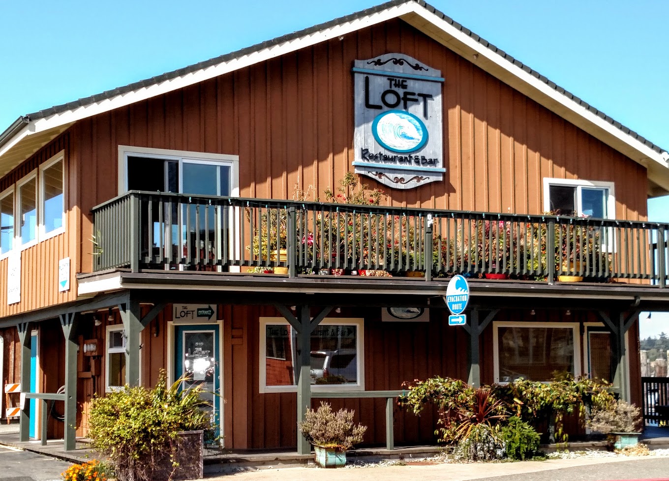 The outside of The Loft Restaurant And Bar. It's a brown building with a patio and potted plants outside. The sign is gray and blue and has a beachy vibe.