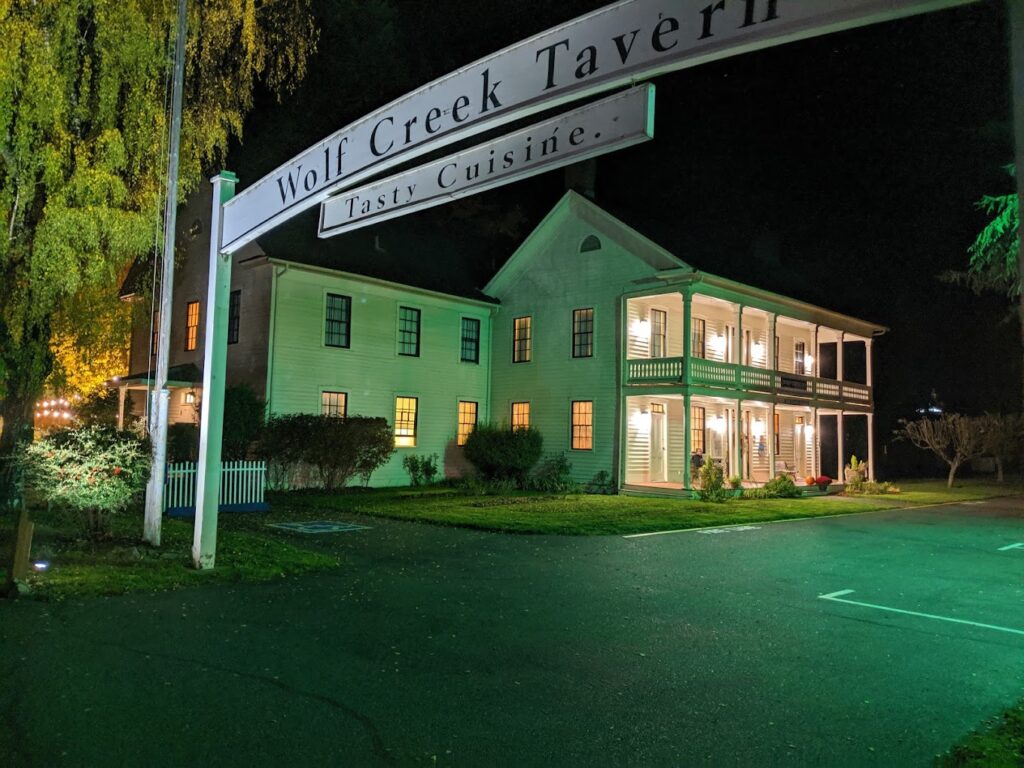 The outside of the Wolf Creek Inn at night. The white Wolf Creek Tavern sign stretches over the street in front of the building. The photo has a green tint and green lighting, giving the entire place a creepy vibe.