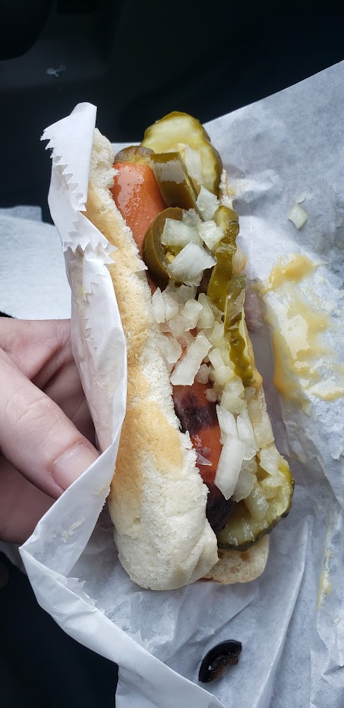 A hot dog in a bun with pickles, onions, and jalapenos held together in a paper food wrapper.