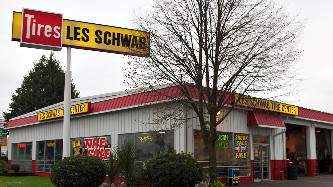 The exterior of a Les Schwab tire store. The building is white with a red roof, and a yellow sign.