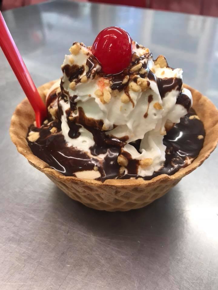 An ice cream sundae with a cherry on top, nuts and a chocolate drizzle. A red spoon is in the sundae.