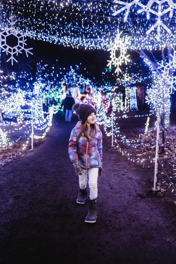 A young girl who is bundled up in warm clothes walks down a path lighted all around with bright Christmas lights at night time.