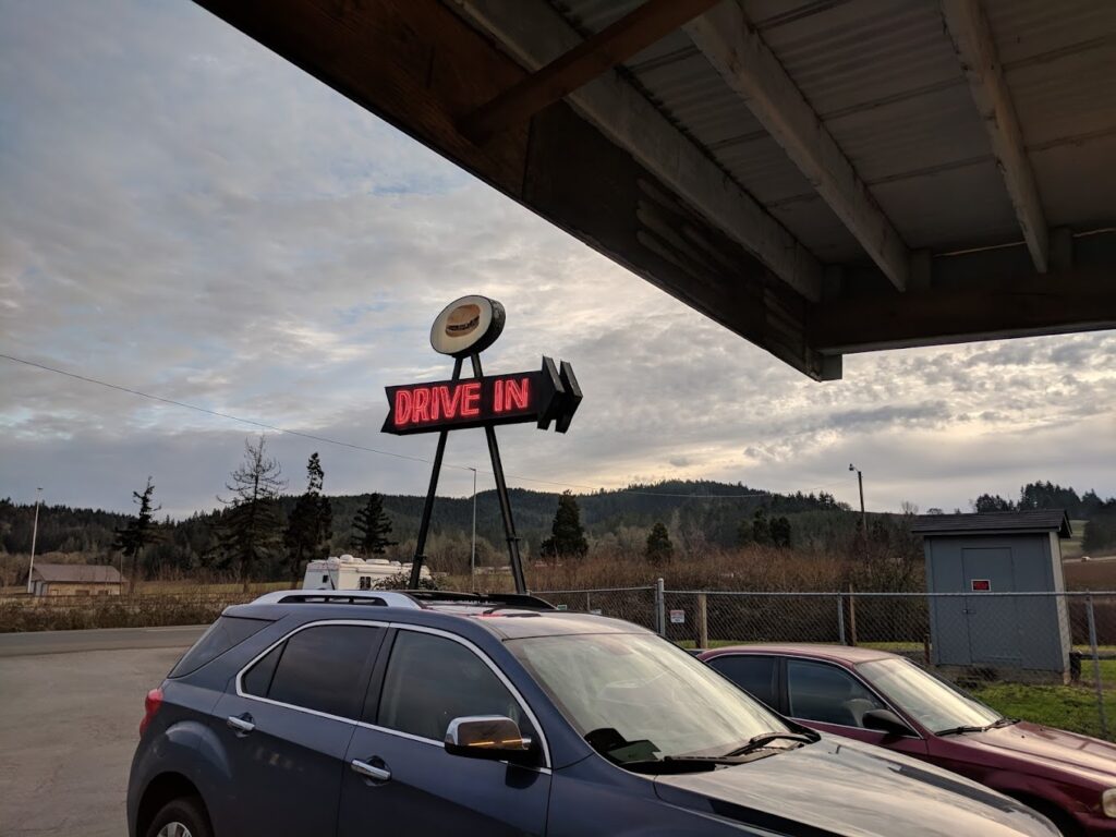 The Drive-In Sign lit up in the day time above two cars in the parking lot.