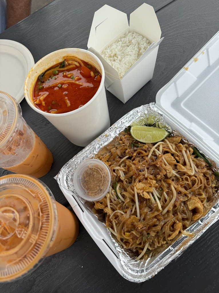 A white Styrofoam box filled with delicious looking Thai food. There are other containers of Thai food and cups of Thai tea beside it.