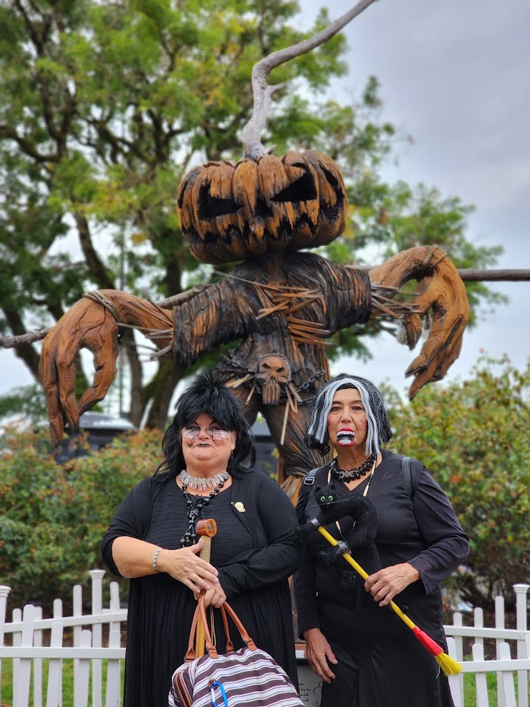 Two people stand in front of a large, spooky carving of a pumpkin head scarecrow.