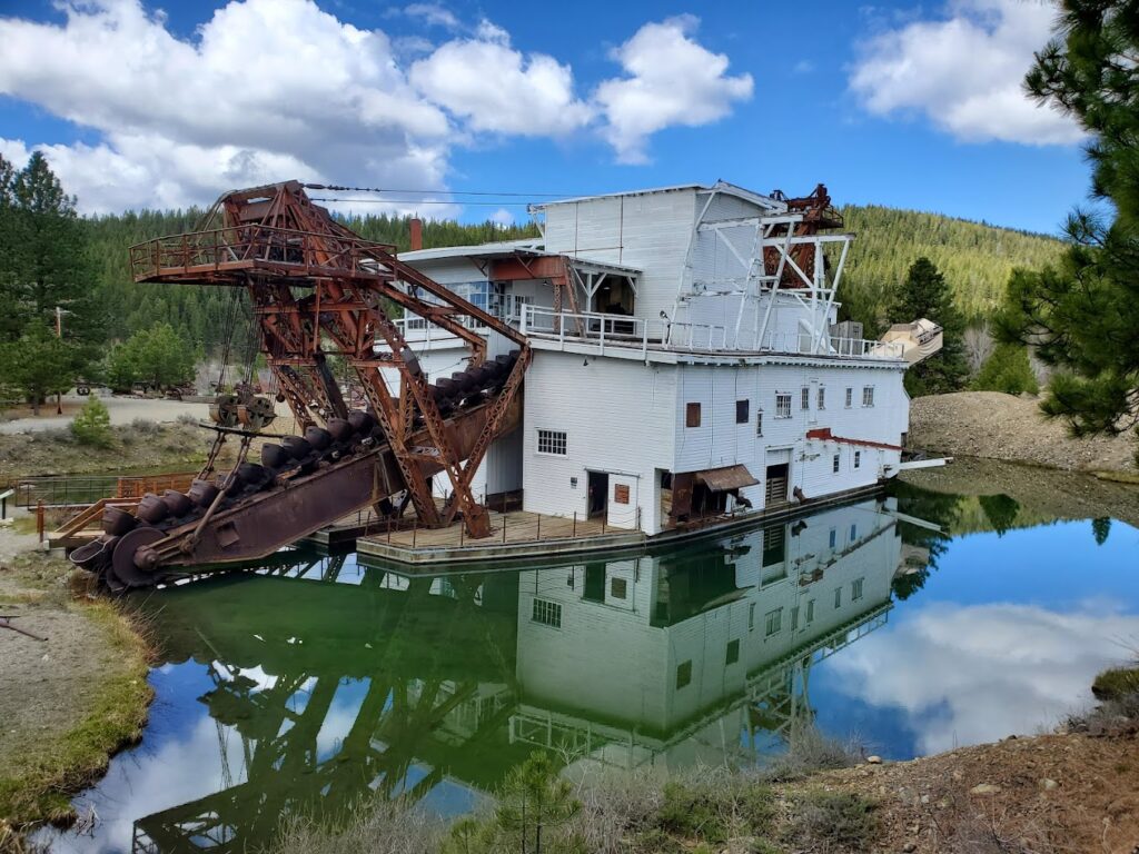 The historic Sumpter Valley Dredge floating in shallow green water. The dredge is painted white and has a huge rusted metal arm.