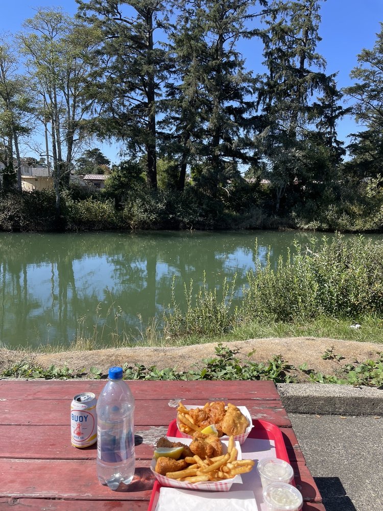 Food on a red picnic table by the river. There are tall evergreen trees on the opposite side of the river.