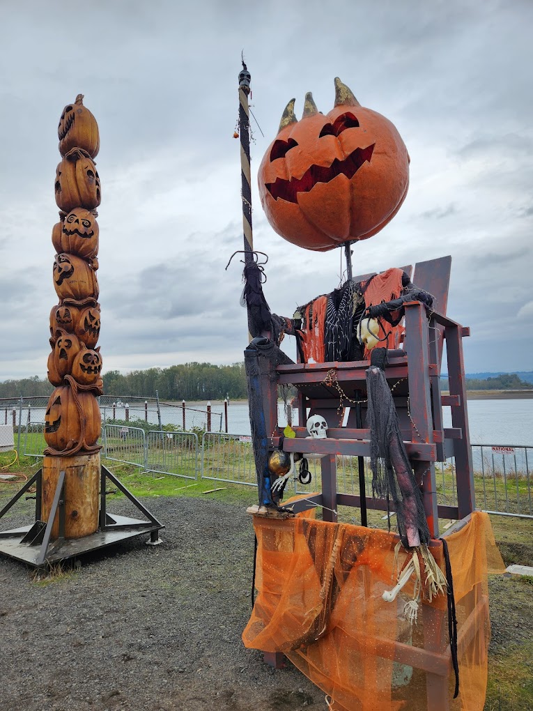 A large pumpkin headed scarecrow sits on a massive wooden chair next to a carved wooden totem pole made of jack-o-lanterns.