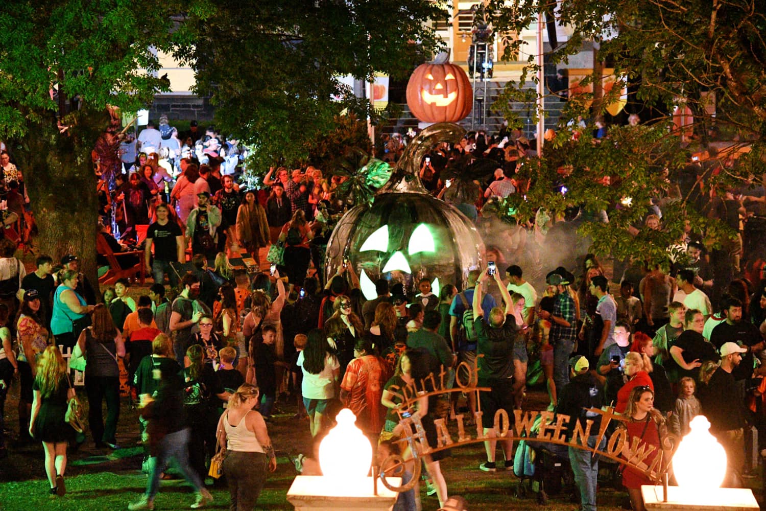 A crowd gathers in front of a massive pumpkin with green glowing eyes at night.