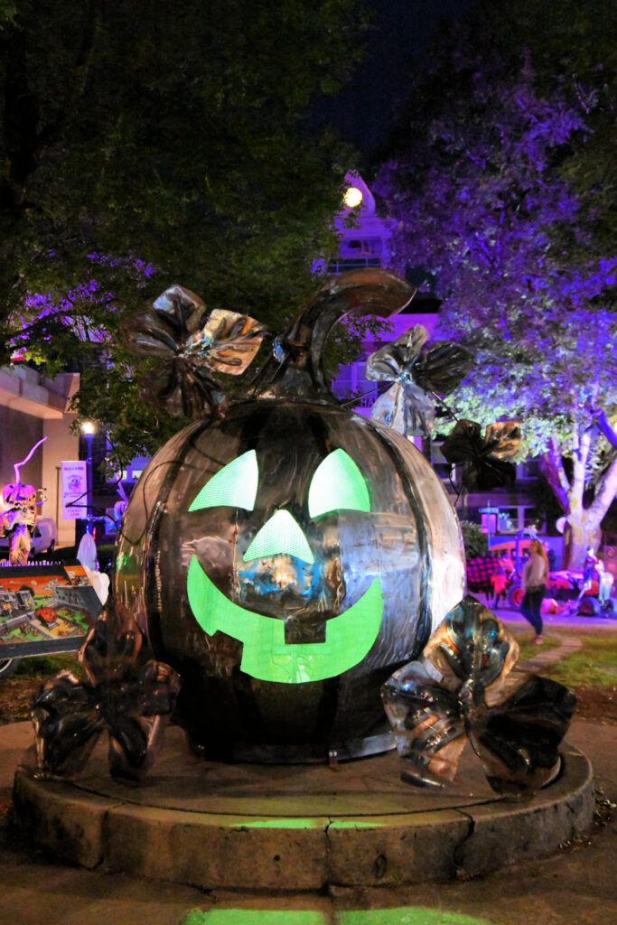 A large metal pumpkin lit up at night. There are purple lights lighting up the trees and landscape behind it.