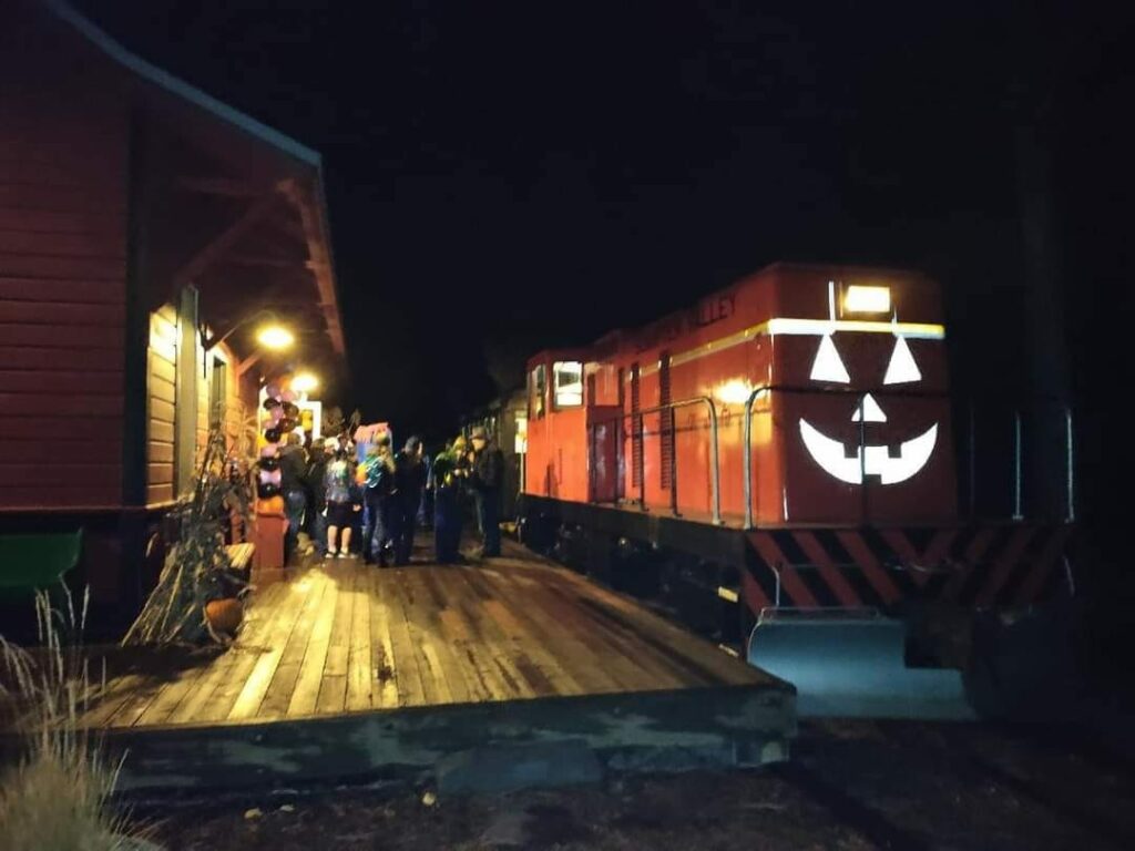 An orange train painted to look like a jack-o-lantern all lit up at night as it sits at the train station.